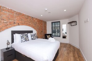 A modern bedroom with a brick accent wall, a large bed with white linens, a nightstand, a mirror, and a window with blinds. - Wood Street Apartments (Liverpool)