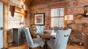 A cozy cabin interior with a wooden interior, a round glass table, two upholstered chairs, a cabinet, a framed picture, and a floor lamp.