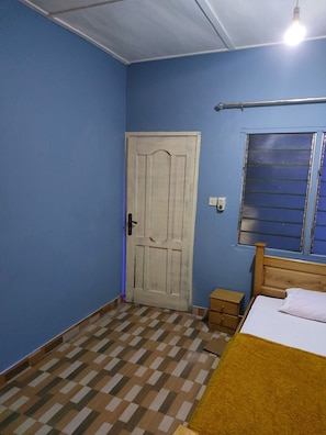 A bedroom with a checkered floor, a wooden bed, a small bedside table, a window with blinds, and a door.