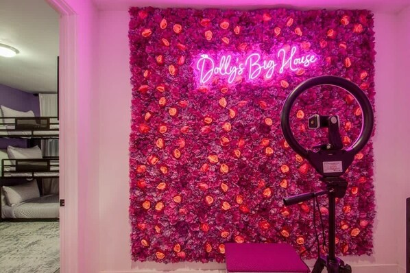 Una habitación con un llamativo mural floral, un letrero de neón que dice "Dolly's Big House" y una luz de aro sobre un trípode.