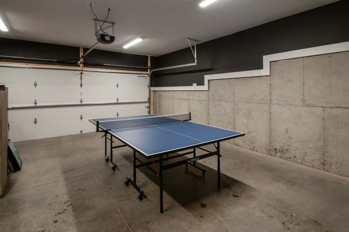 Une table de ping-pong se trouve dans une pièce dont le sol et les murs sont en béton, avec un projecteur fixé au plafond et une porte blanche dotée d’une poignée en métal.