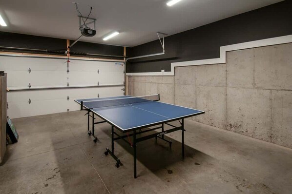 Una mesa de ping pong en una habitación con suelo de concreto y paredes, un proyector montado en el techo y una puerta blanca con manija de metal.