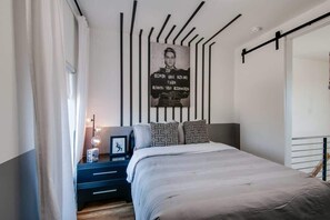Un dormitorio con una cama, una mesita de noche con lámpara, un cartel en la pared y una ventana con cortinas.