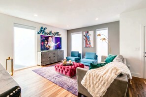 Ein modernes Wohnzimmer mit einem großen Flachbildfernseher, einem rot gepolsterten Ottomanen, einem grauen Sofa mit weißer Decke und einem blauen Sessel.