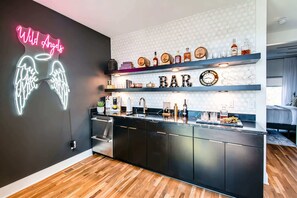 A modern kitchen with a neon sign, wooden flooring, and a bar area with shelves of bottles and glasses.