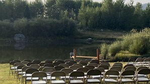 Rows of chairs arranged in a semi-circle facing a body of water, with trees and a grassy area in the background.