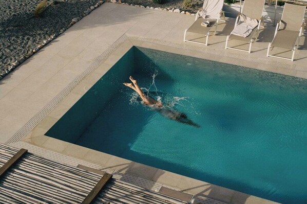 A rectangular swimming pool with a person swimming, surrounded by a tiled deck and lounge chairs.