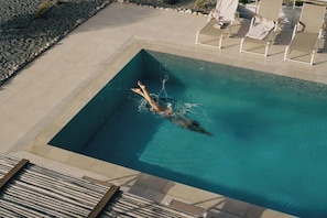Una piscina rectangular con una persona nadando, rodeada de una terraza con baldosas y sillas de descanso.