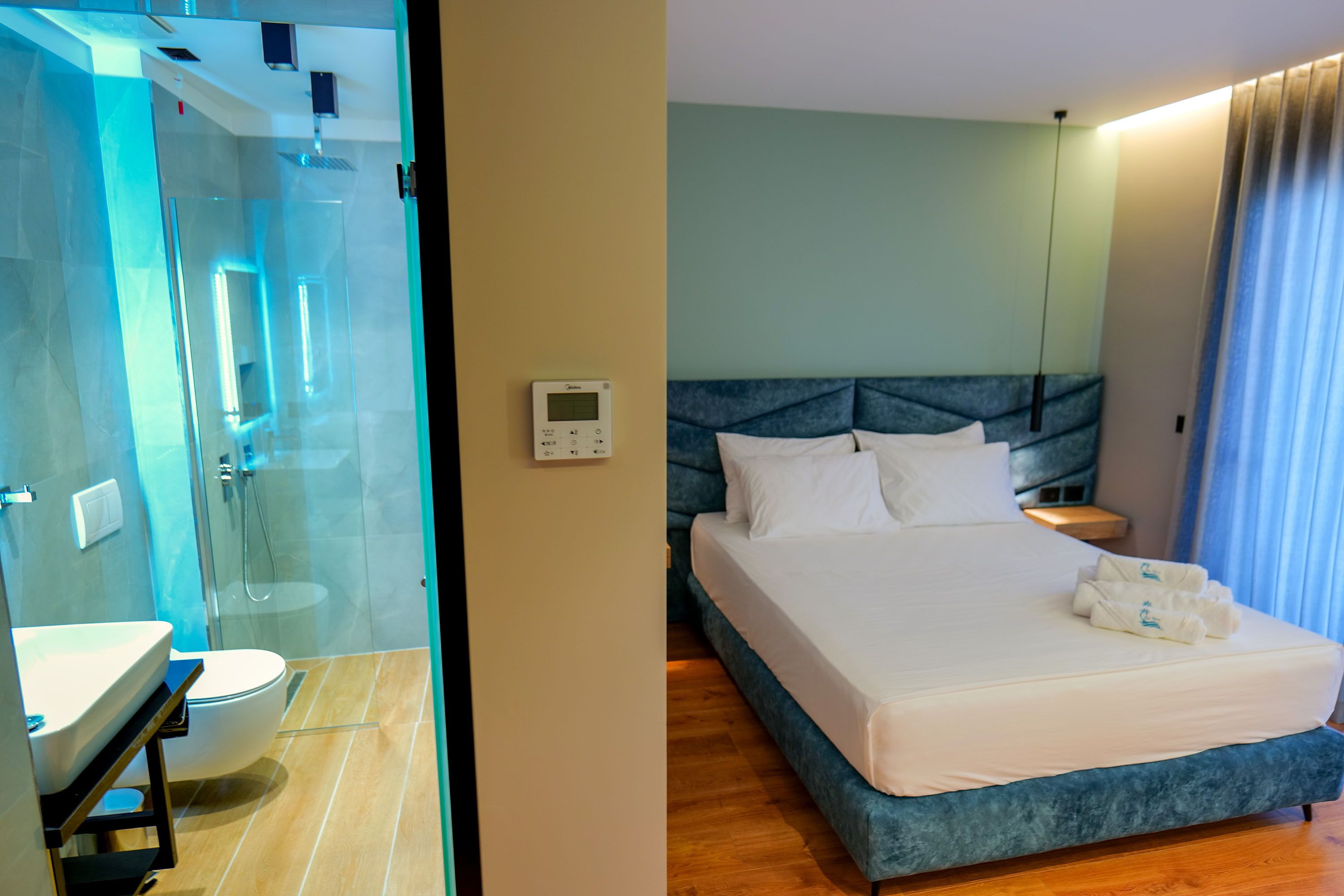 A hotel room with a glass-enclosed shower, a bed with white linens, and a control panel on the wall.