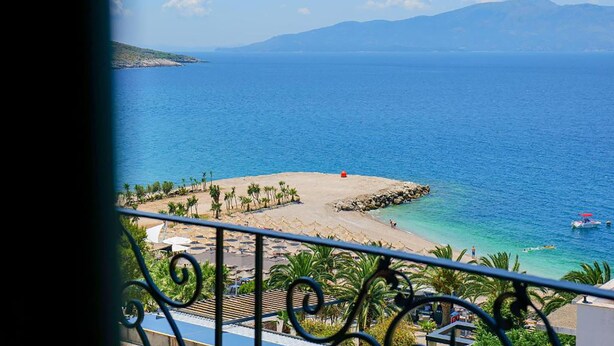A balcony with a view of a beach, clear blue sea, and distant mountains. - Seawave Hotel (Sarandë)