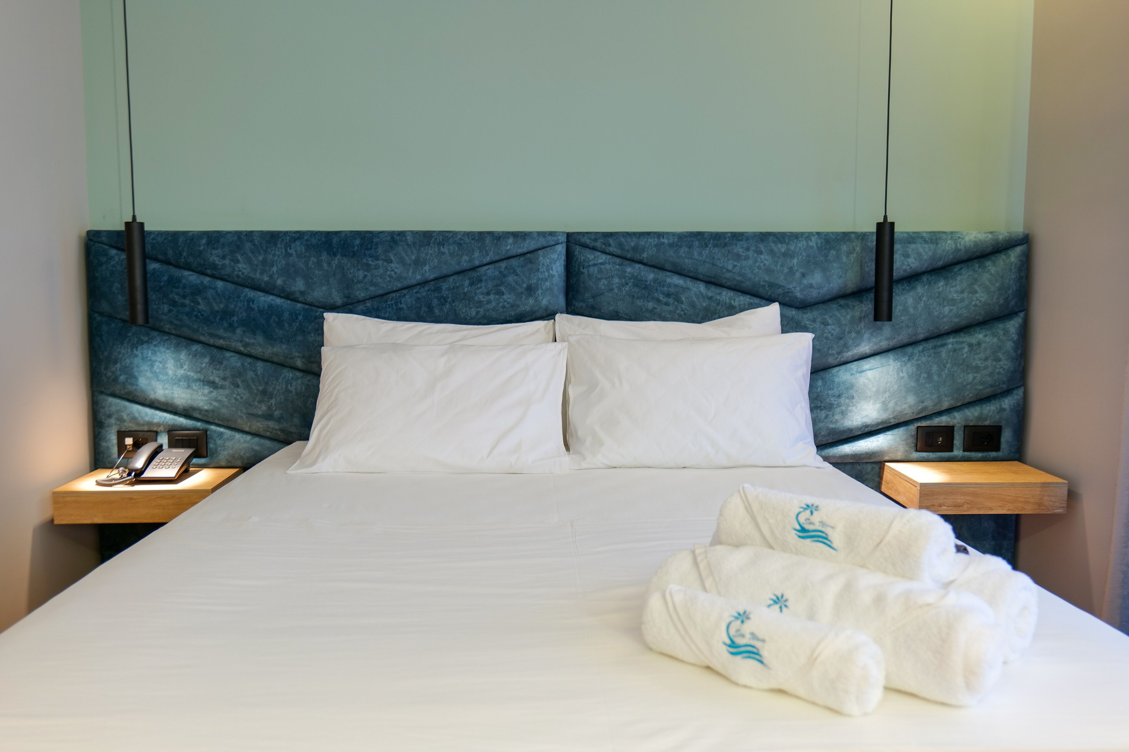 A neatly made bed with white linens, a headboard with a geometric pattern, and two bedside tables with a phone and a lamp.