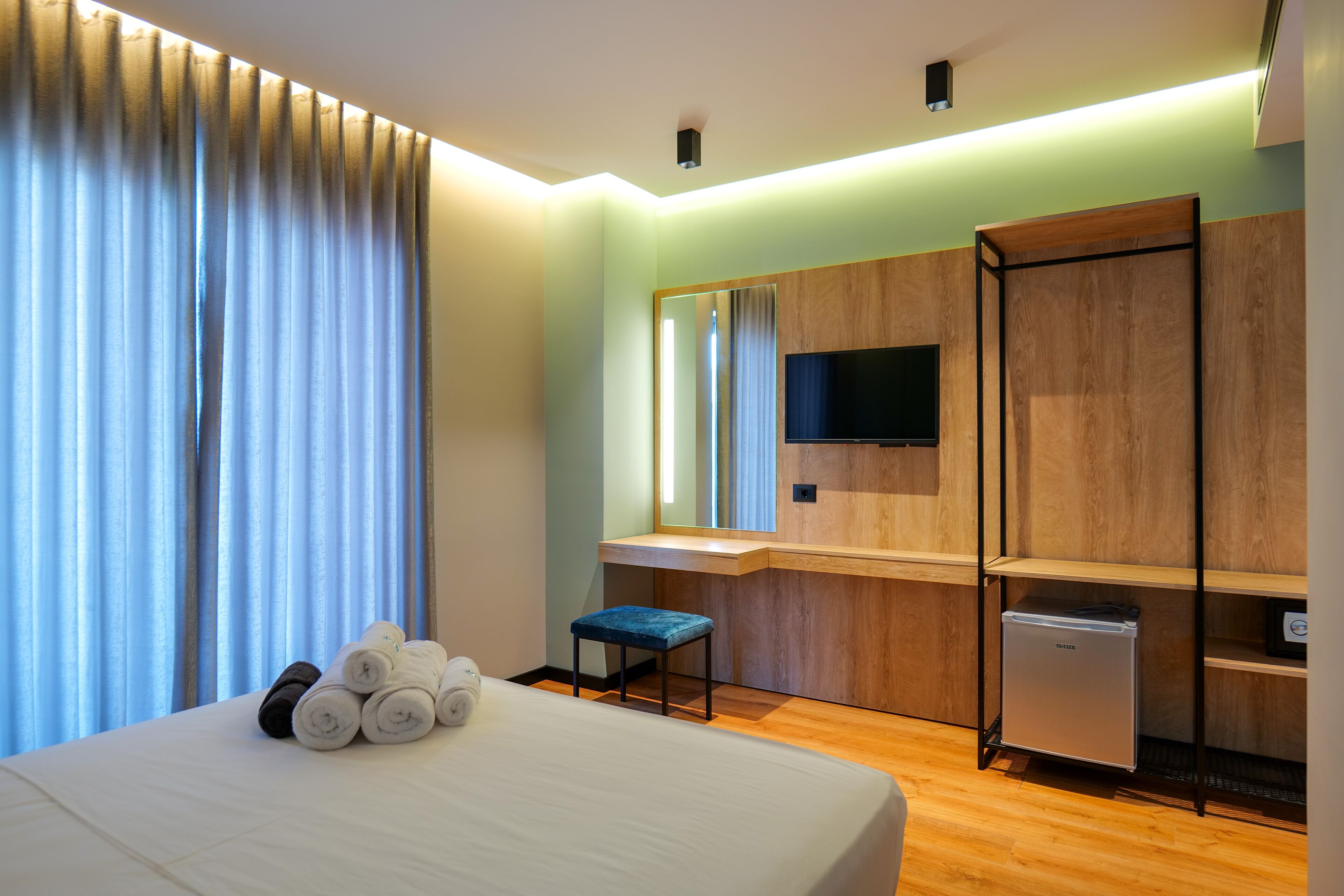 Ein Hotelzimmer mit Bett, einem kleinen Tisch mit Handtüchern, einer Bank, einem Spiegel, einem Fernseher und einer Minibar.
