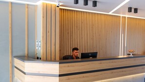A reception desk with a wooden panel backdrop, a computer, and a vase with flowers. - Seawave Hotel (Sarandë)