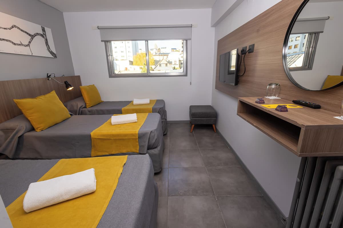 A hotel room with two beds, a small desk, a mirror, and a window with a view of buildings.