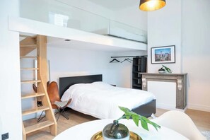 A modern bedroom with a lofted sleeping area, a bed with white bedding, a chair, a small table with a plant, and a fireplace. - Amazing studio located in Ghent (Gent)