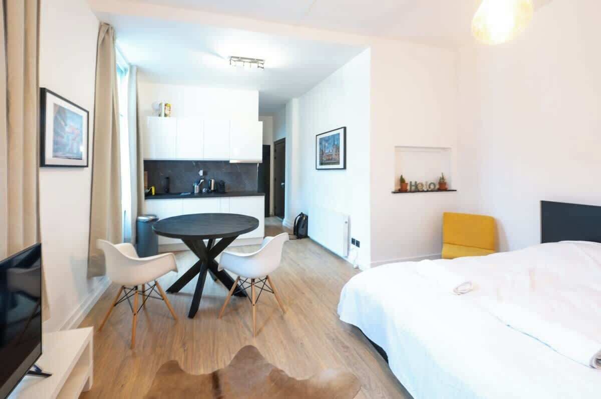 Een moderne hotelkamer met een bed, een ronde eettafel met stoelen, een klein keukentje en een gele fauteuil.