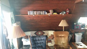 Une pièce chaleureuse avec un plafond en bois, une bibliothèque remplie de divers objets, un canapé agrémenté d’un plaid à motifs, deux fauteuils, une lampe et une petite table sur laquelle se trouve un four à micro-ondes.