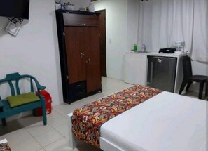 Een kleine, eenvoudige kamer met een bed, een bureau, een kledingkast en een stoel.