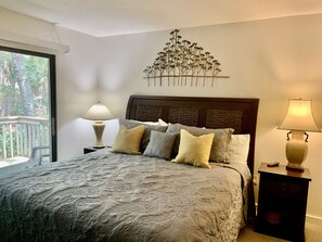 A bedroom with a large bed, two bedside tables, a decorative wall piece, and a view of trees through a window.