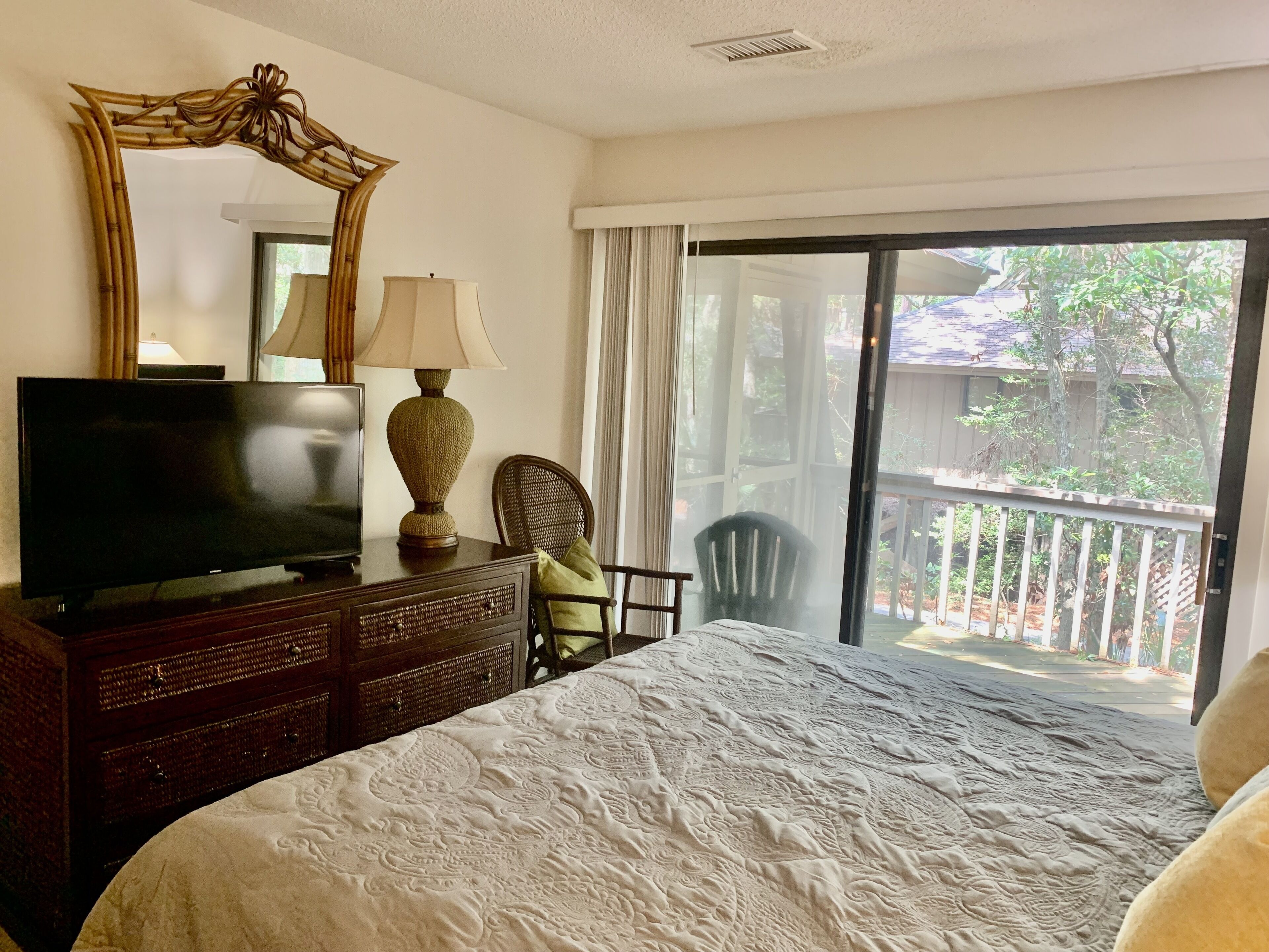 Un dormitorio con una cama grande, un televisor sobre una cómoda, una silla, una lámpara y un balcón con vista a árboles.