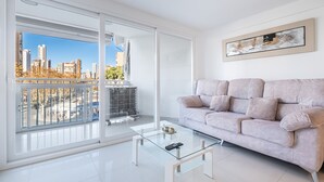 Un salon moderne avec une table basse en verre, un canapé de couleur claire et une vue sur la ville grâce à de grandes fenêtres.