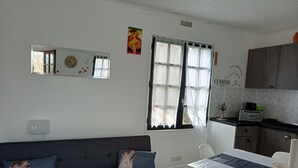A modern kitchen with a dining area, a gray sofa with decorative pillows, and a window with sheer curtains. - Maison ds Marais Poitevin (Taugon)