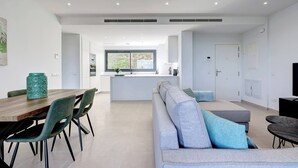 A modern living room with a sofa, dining table, and chairs. There is a kitchen area with a window and a door. - Modern 2 Bed Apartment in Trendy La Cala Golf Resort (Mijas)