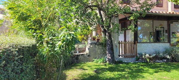Un edificio rústico con pared de piedra, un portón de madera y un pequeño jardín con diversas plantas y un árbol.