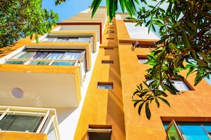 A multi-story building with balconies, large windows, and a tree with green leaves in the foreground.