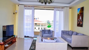 A living room with a sofa, a TV, and a view of buildings outside. - CherryWood Apartments (Nairobi)