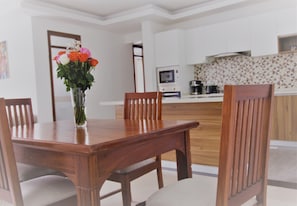 A dining area with a wooden table and chairs, a vase with flowers, and a kitchen in the background.