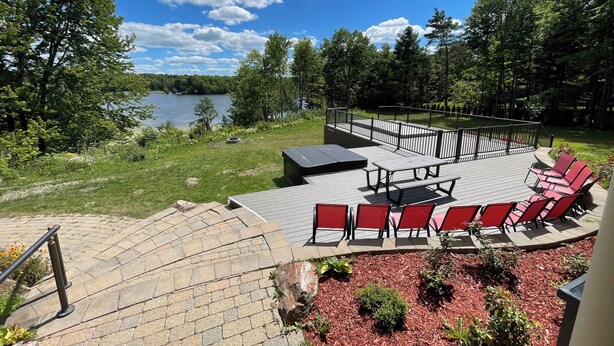 A deck with red chairs and a table overlooking a lake, surrounded by trees and a grassy area. - Cozy cottage home close to nature includes game room, fitness equip, & Hot Tub. (Portland)