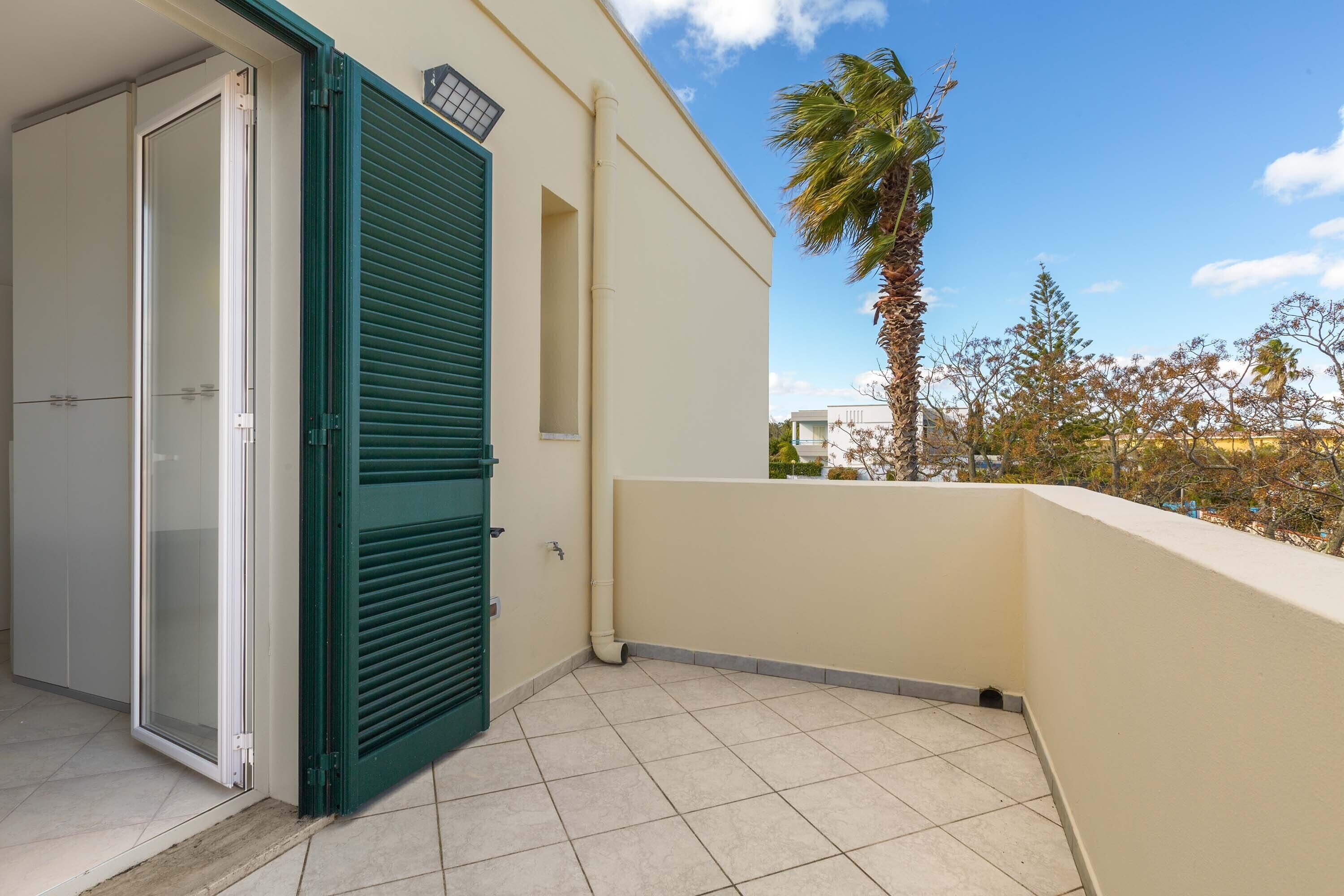 A balcony with a tiled floor, a green door, and a view of trees and buildings.