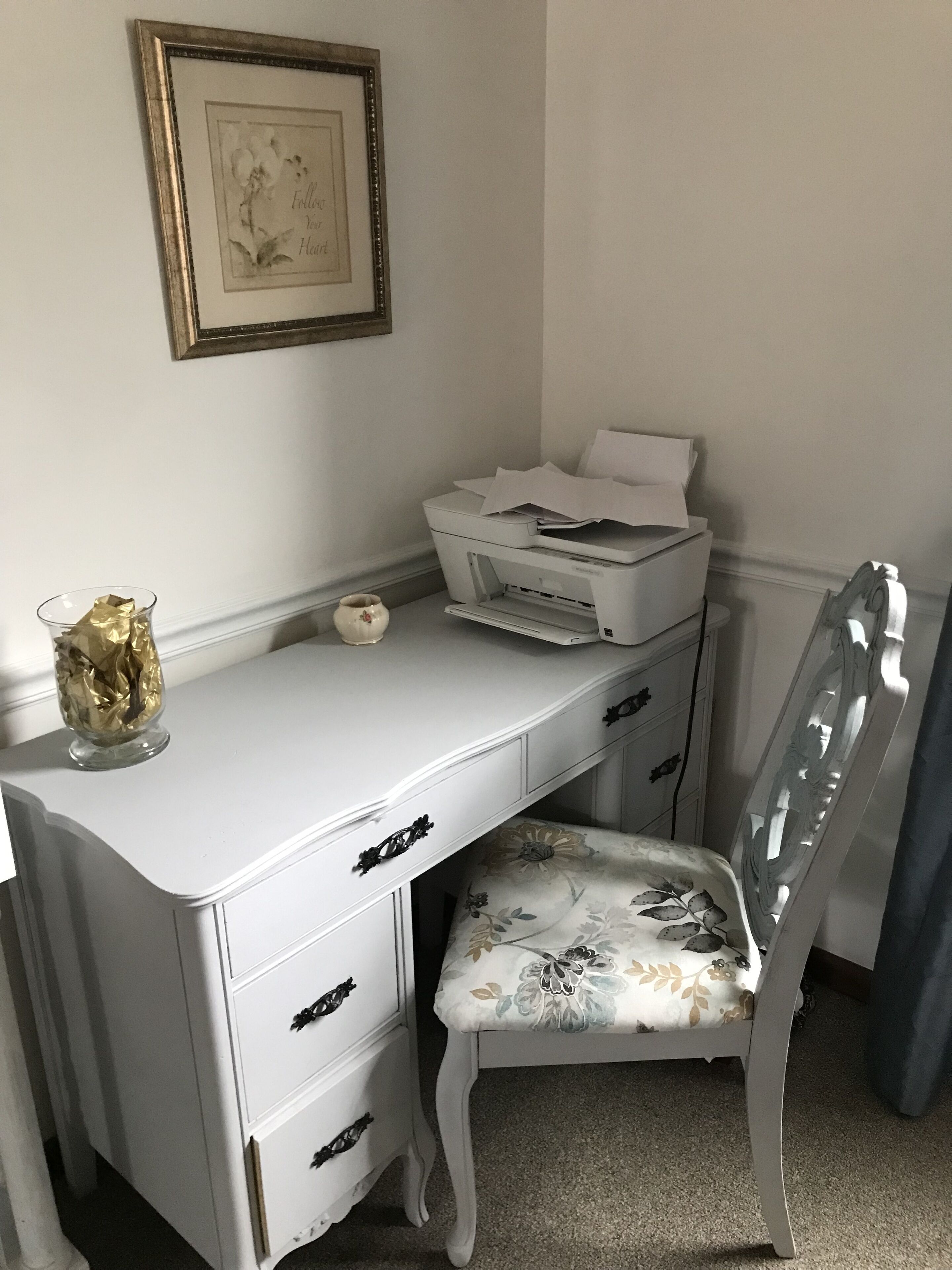 Ein weißer Schreibtisch mit Schubladen und einem Drucker, ein Stuhl mit floralem Bezug, ein gerahmtes Bild an der Wand und eine Vase mit einem goldenen Gegenstand auf dem Schreibtisch.
