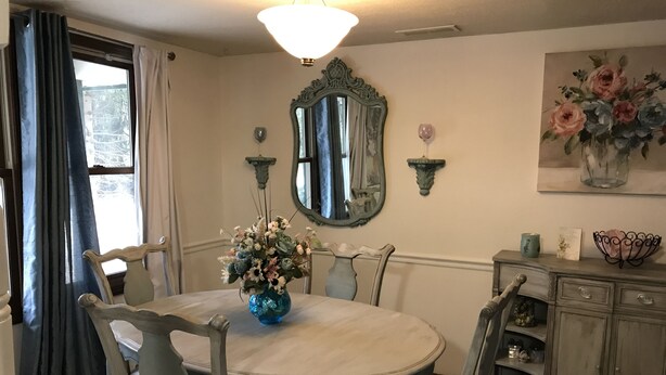 A dining room with a round table, chairs, a chandelier, a mirror, and a floral painting.