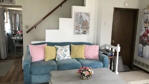 A living room with a blue sofa, a white coffee table with a floral centerpiece, a staircase, and a door with a 'Welcome' sign.