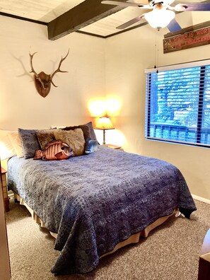 Un dormitorio con cama, lámpara, ventana con persianas, ventilador de techo y cabeza de venado colgada en la pared.