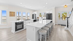 A modern kitchen with a large island, bar stools, and a view of the beach. - Fall Available | Oceanfront Home w Panoramic Views (Ocean Isle Beach)