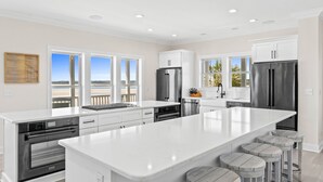 A modern kitchen with a large island, stainless steel appliances, and a view of the beach. - Fall Available | Oceanfront Home w Panoramic Views (Ocean Isle Beach)