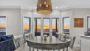 A dining area with a large table and chairs, a hanging light fixture, and large windows offering a view of the sunset over the water. - Fall Available | Oceanfront Home w Panoramic Views (Ocean Isle Beach)
