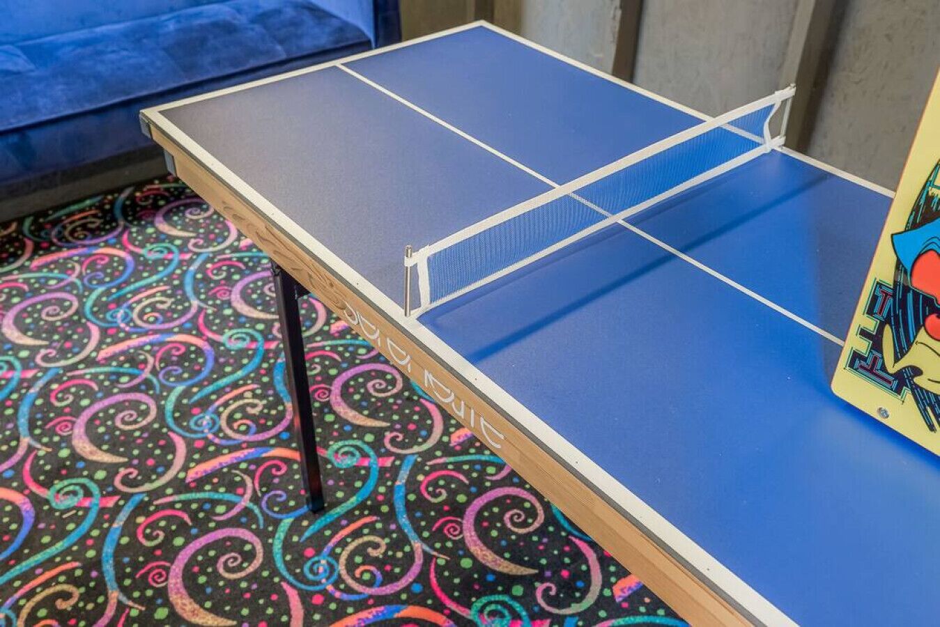 A ping pong table with a blue playing surface and a wooden frame, placed on a patterned carpet.
