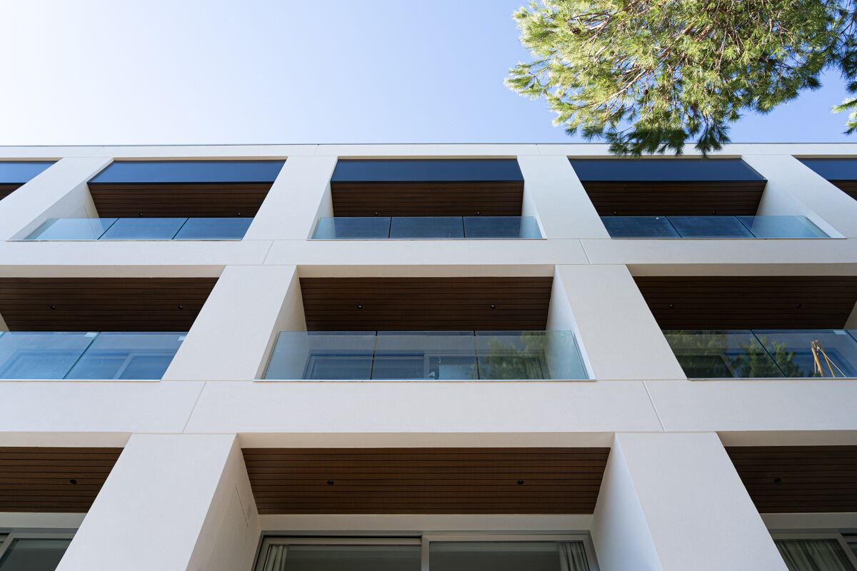 Um edifício moderno com vários andares, janelas amplas e uma árvore visível ao fundo.