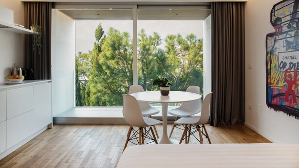 A modern room with a large window, a round table with chairs, a bed, and a painting on the wall.