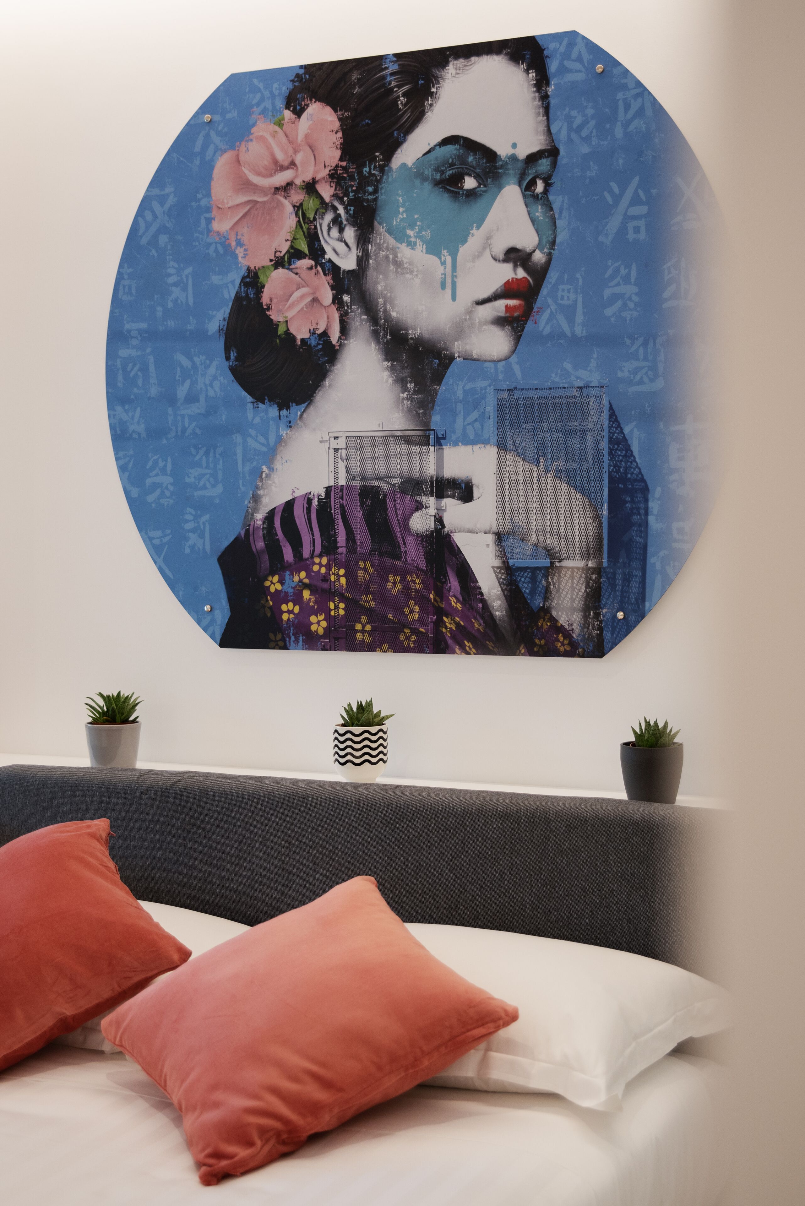 A wall-mounted artwork depicting a woman with flowers, a bed with red and white pillows, and two potted plants.