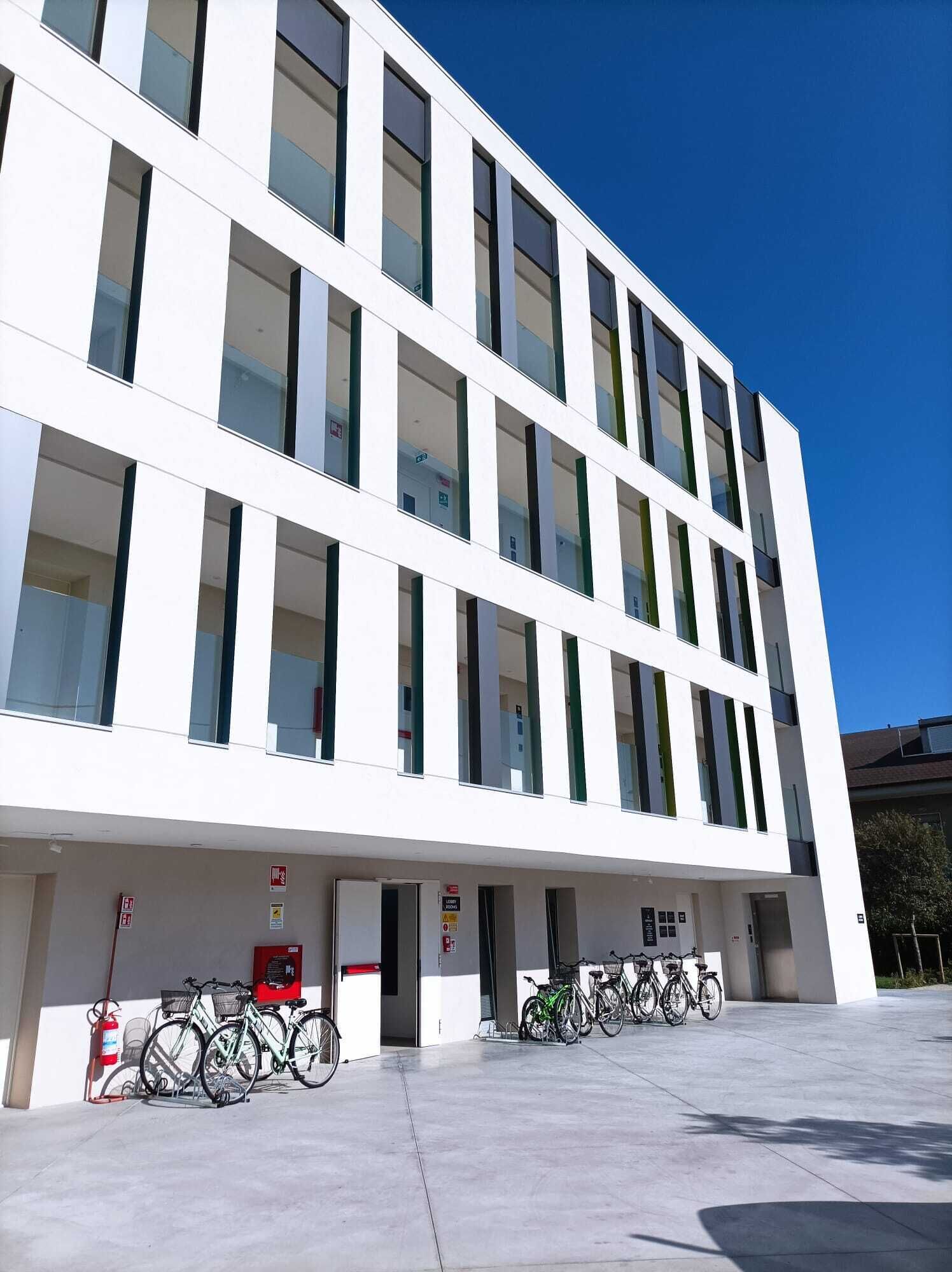A modern building with a white facade, multiple windows, and a covered entrance area with bicycles parked outside.