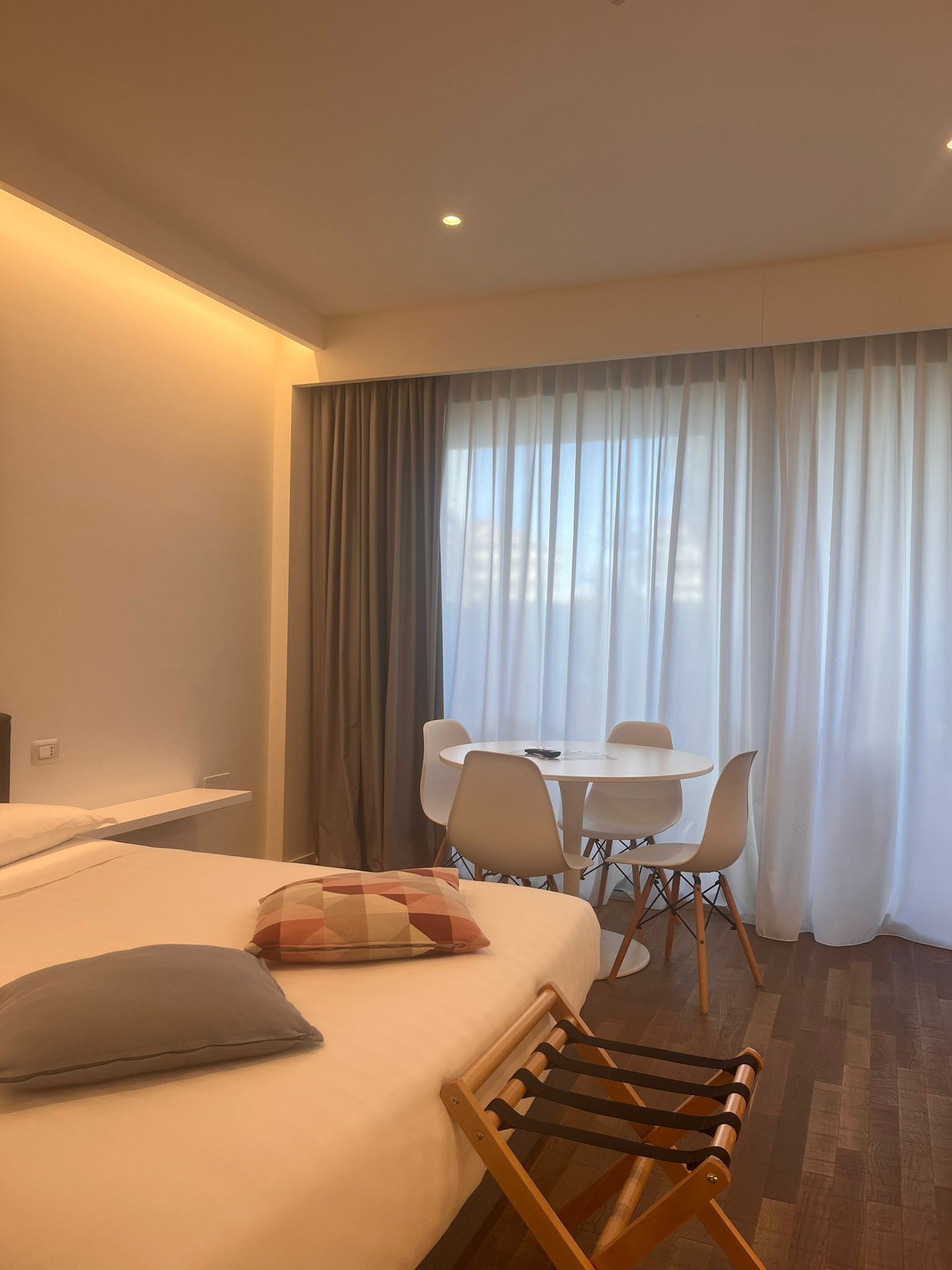 Habitación de hotel con cama, una silla de madera, una mesita y vistas al exterior a través de cortinas traslúcidas.
