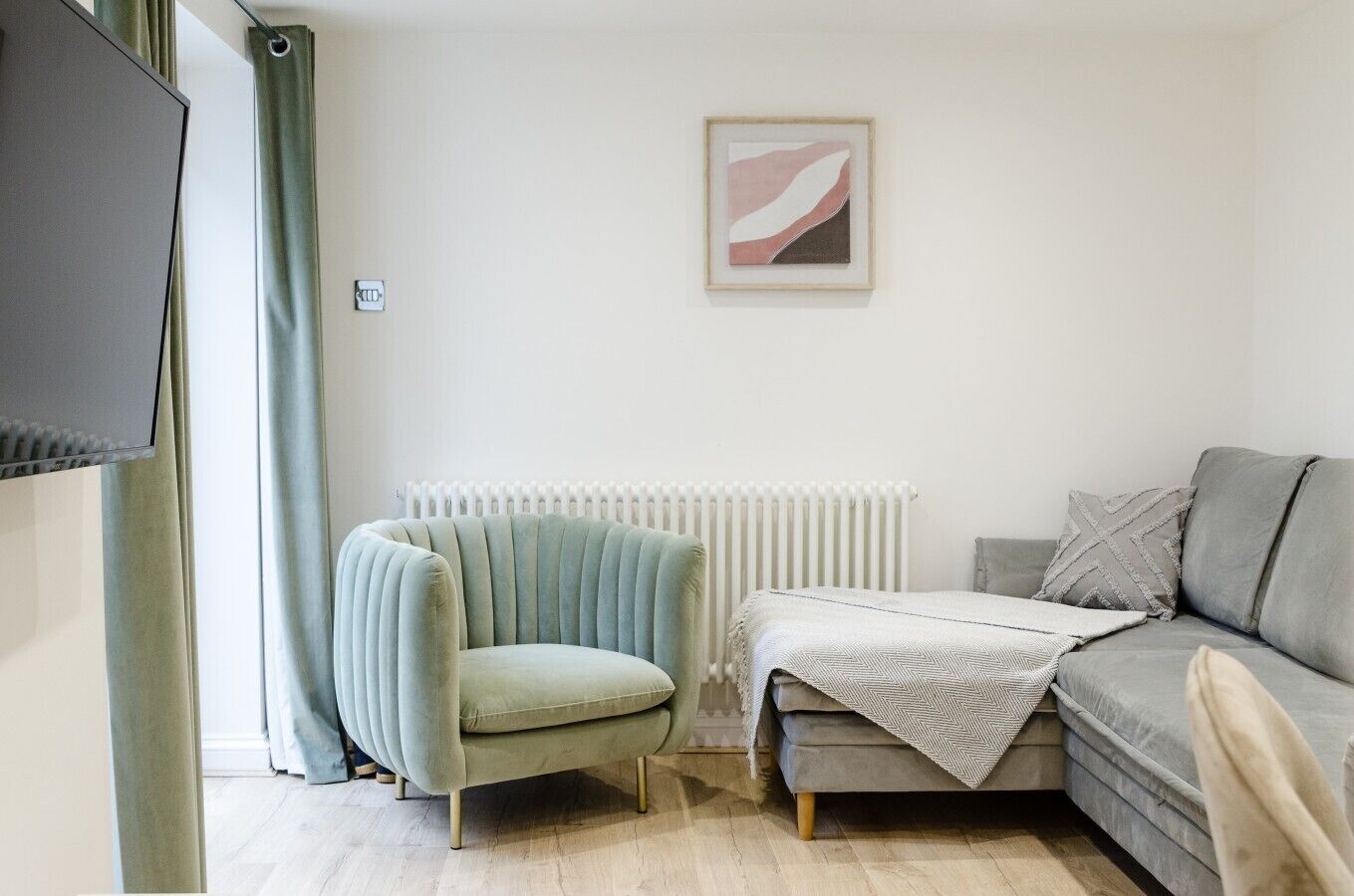 Un salon avec un fauteuil vert clair, un canapé gris, un radiateur blanc et un tableau abstrait encadré accroché au mur.