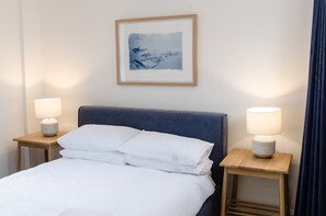 Habitación de hotel con cama, dos mesitas de noche con lámparas, un cuadro enmarcado en la pared y una cortina azul.
