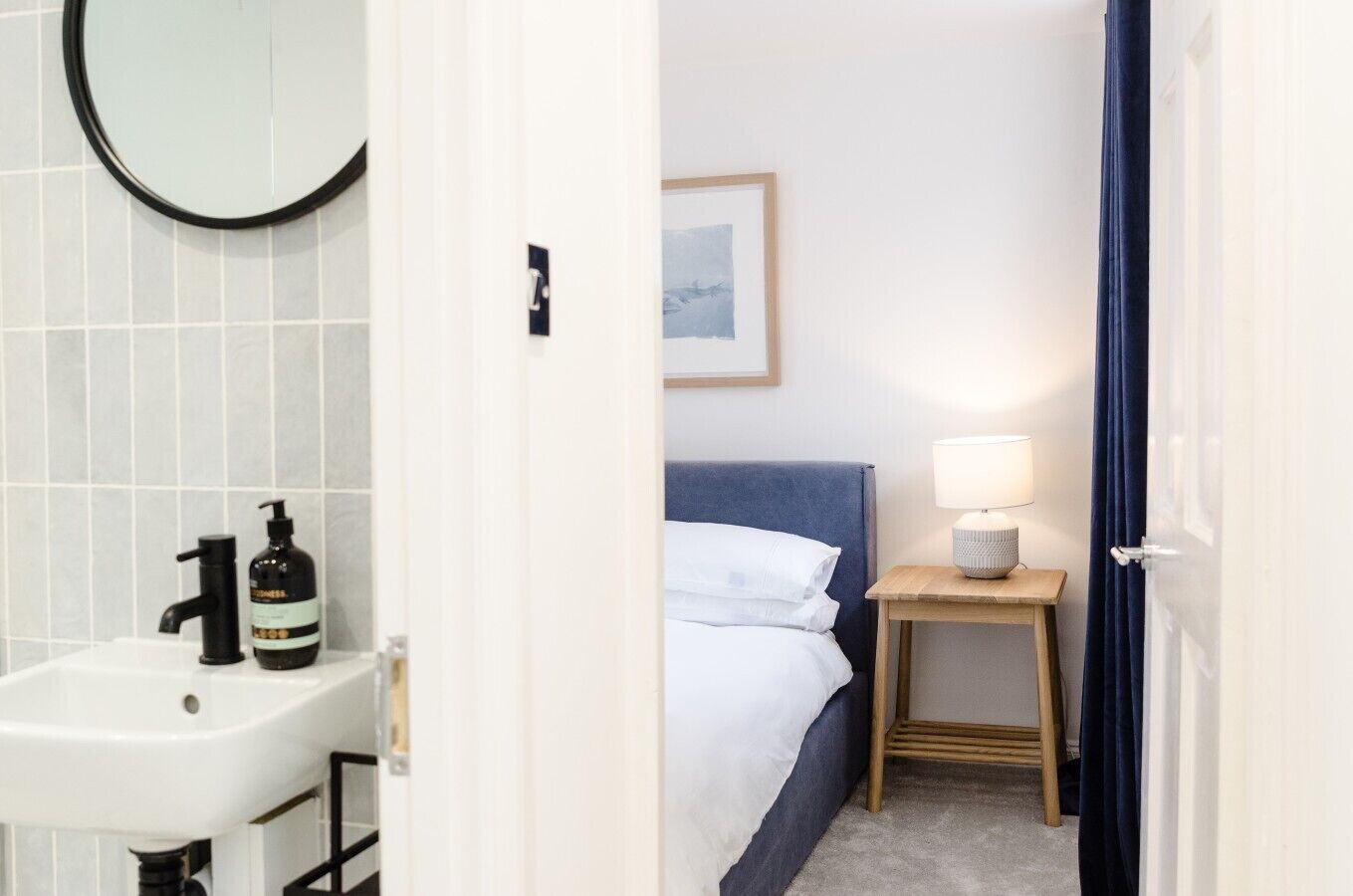 Een hotelkamer met een wit wastafeltje, een bed met witte lakens, een blauw hoofdeinde, een nachtkastje met een lamp, en een ingelijst schilderij aan de muur.