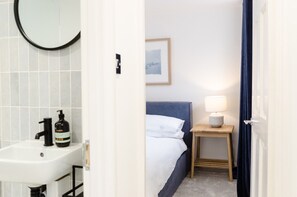 Une chambre d’hôtel avec un lavabo blanc, un lit avec du linge de lit blanc, une tête de lit bleue, une table de chevet avec une lampe et un tableau encadré au mur.